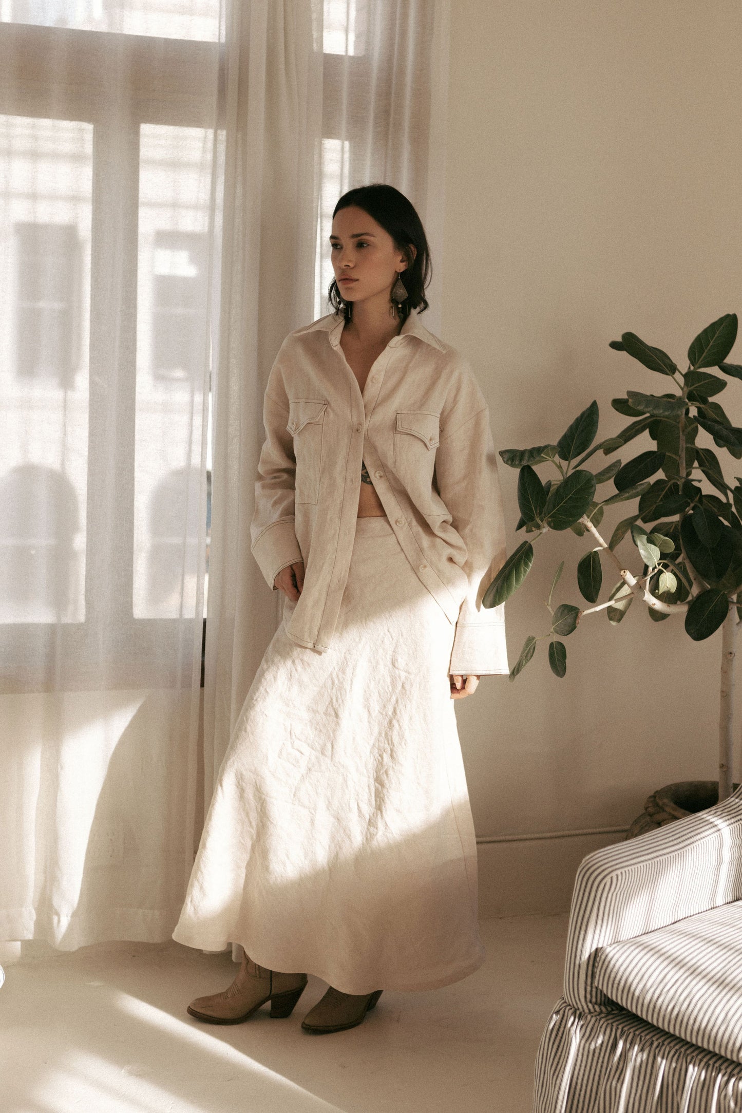 Woman in a beige shirt and white skirt standing in a bright room with curtains and a plant.