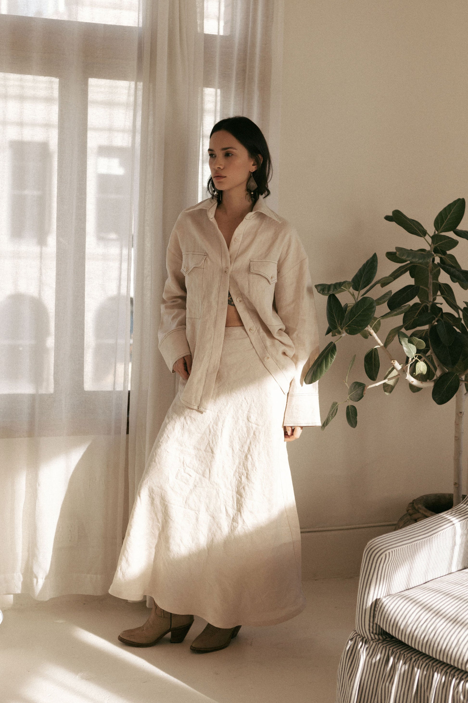 Woman in a beige shirt and white skirt standing in a bright room with curtains and a plant.